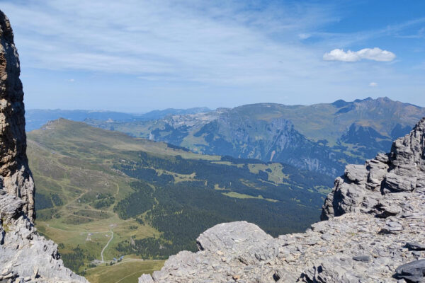 Ausblick mit blauem Himmel und einigen Wolken auf grüne Bergwiesen und Wälder.