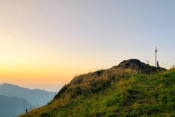 Abendrot hinter den Bergen und Gipfelkreuz mit Person nahe der Alp Oberfeld